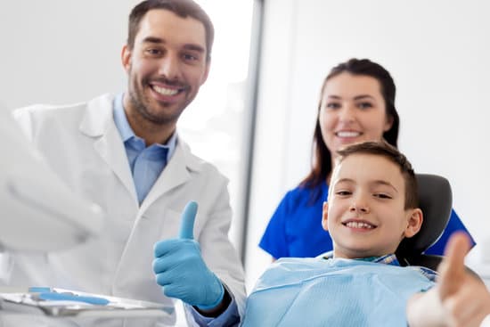 A kid on the dental chair showing thumbs up with a smiling face. The Pediatric dentist and the mom of the kid are seen as well. All smiling