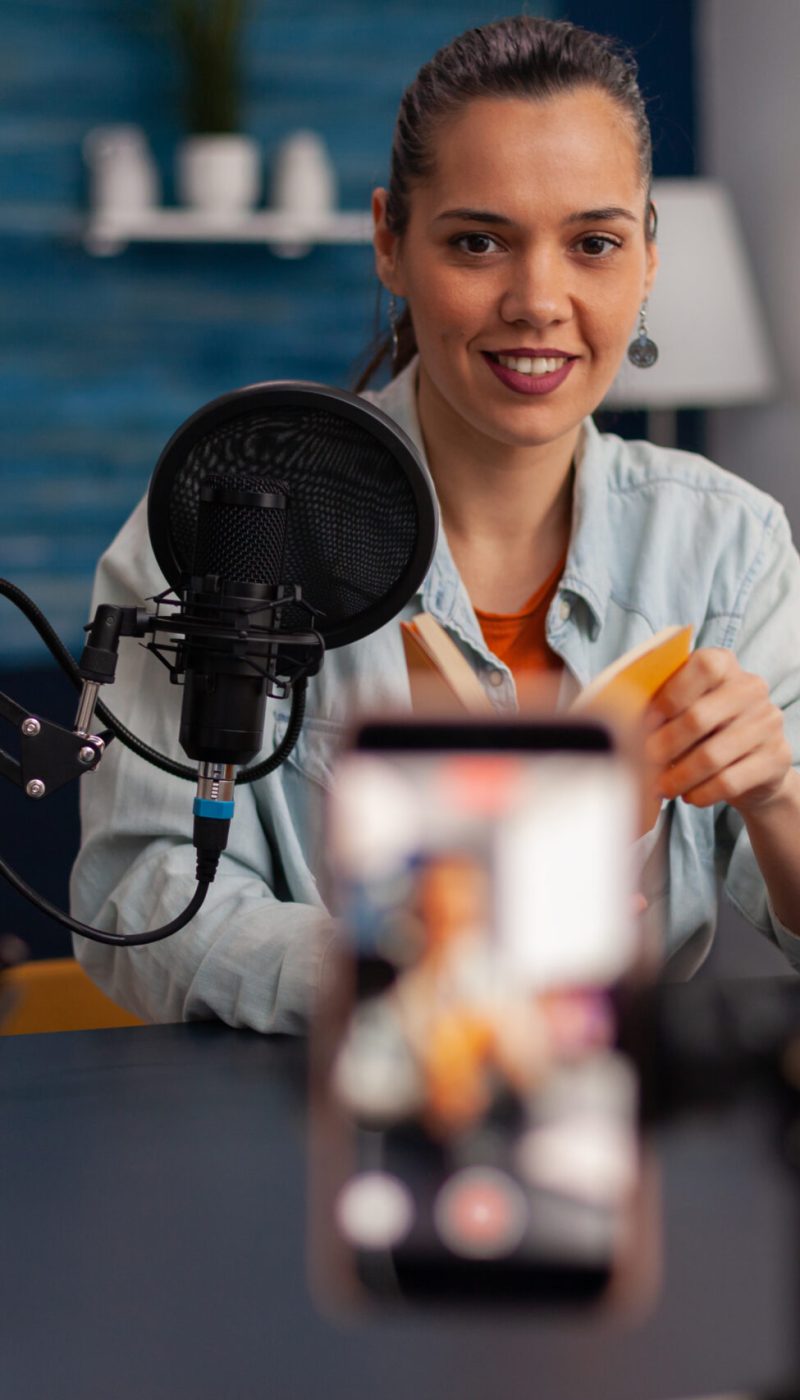 Influencer recording yellow book review for social media channel. Woman content creator presenting product recommendation for online vlog in home studio with microphone and laptop.